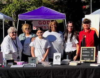 A group of six women stands behind a table displaying books, journals, and bundles for sale at an outdoor event. Bright sunlight and a purple canopy add to the vibrant atmosphere.
