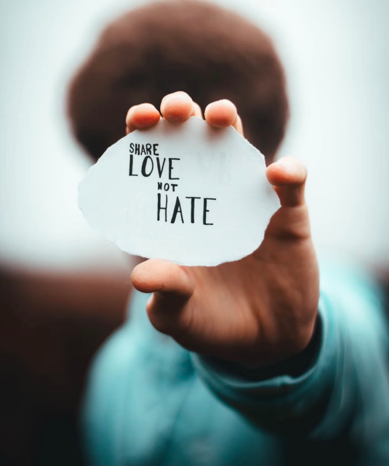 A hand holds a torn paper with the message "Share Love Not Hate," promoting positivity and compassion. The blurred background emphasizes the message.