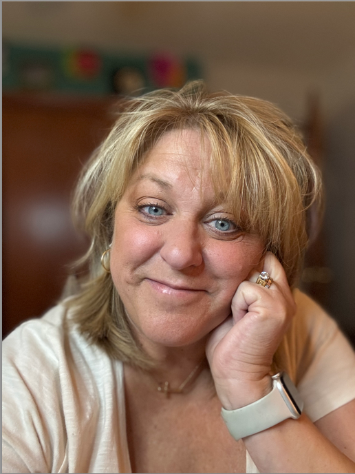 Smiling woman with blonde hair holds her chin in her hand, showcasing her engagement and warmth. Background is softly blurred, suggesting a cozy indoor setting.