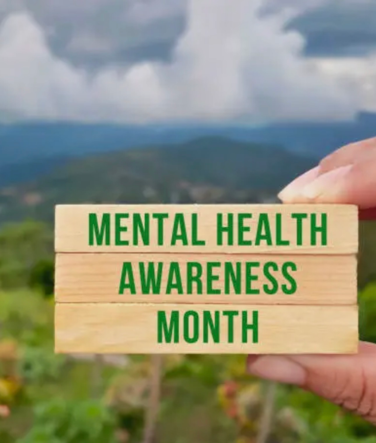 Hands hold three wooden blocks stacked vertically, displaying "Mental Health Awareness Month" in green text. Cloudy sky and nature in the background.